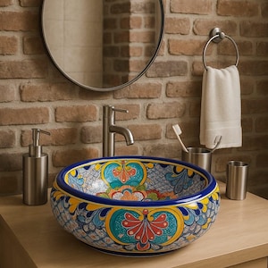 May include: A decorative, hand-painted ceramic vessel sink with a vibrant floral design in blue, yellow, and red. The sink is set on a light wood vanity with a stainless steel faucet and accessories, against a brick wall.