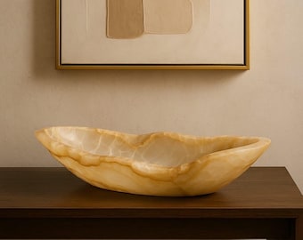 May include: A decorative, oblong bowl made of light-colored onyx. The bowl has a natural, organic shape with a smooth, polished surface. The onyx has a translucent quality, with variations in color from cream to light brown.