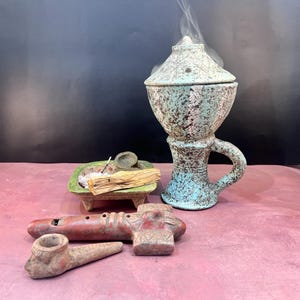 May include: A teal and brown ceramic incense burner with a handle, a small wooden incense stick, and a small ceramic incense holder with a wooden incense stick on a red surface.