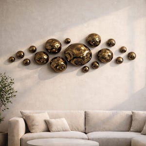 May include: A wall art installation featuring a collection of bronze-colored, irregularly shaped spheres of varying sizes. The spheres are mounted on a neutral-toned wall above a beige sofa, creating a modern, abstract design element for home decor.