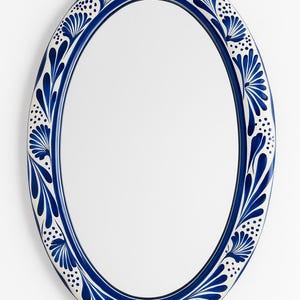 May include: Oval mirror with a blue and white floral design. The frame is a deep blue with a white border featuring a repeating pattern of stylized leaves and dots. The mirror has a reflective surface.