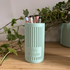 May include: A pale green pencil pot with vertical lines and the name "Mrs Wright" embossed on the front. It holds several pens with black tips and rose gold details. The background features greenery and a wooden surface.