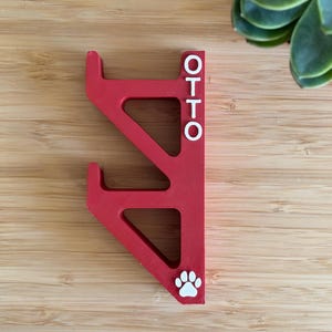 May include: A red wooden phone stand with the name "OTTO" in white lettering. The stand has a paw print at the bottom and is designed to hold a phone. The stand is on a wooden surface.