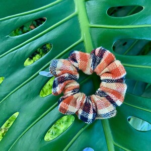 May include: A scrunchie with a plaid pattern in shades of orange, tan, and navy blue. The hair accessory is made of fabric and is circular in shape. The scrunchie is resting on a large, green leaf with holes.