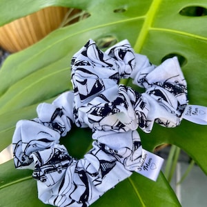 May include: Two white scrunchies with a black and white print of a cartoon character. The scrunchies have a white tag that says "Stitchin' Mom" with a heart.