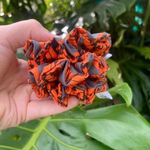 May include: A scrunchie with a repeating pattern of black shapes on a gray background, set against a vibrant orange fabric. The scrunchie is held in a hand, with lush green foliage in the background.