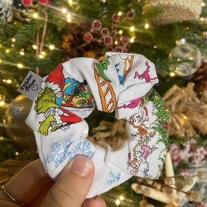 May include: White fabric scrunchie with a Christmas-themed print featuring the Grinch and other characters. The scrunchie is held in a hand, with a Christmas tree and ornaments in the background. The scrunchie has the text "Stitchin' Mom" on it.