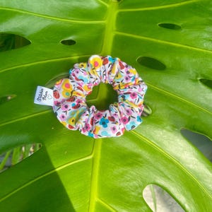 Può includere: Un colorato elastico per capelli con un motivo floreale e di insetti, con accenti rosa, gialli, blu e rossi su sfondo bianco. L'elastico per capelli è in tessuto e ha una piccola etichetta con scritto "Stitchin' Mom".