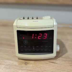 May include: A cream-colored GE digital alarm clock radio with a black display panel showing the time 1:23 in red LED digits. The device features a radio frequency tuner for FM and AM bands, top-mounted control buttons, and a side-mounted dial.