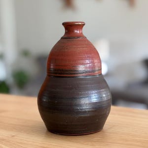 Peut inclure: Un vase en céramique fait à la main, en deux tons. La partie supérieure est brun rougeâtre, tandis que la partie inférieure est brun plus foncé. Le vase a une base arrondie et un col étroit, posé sur une surface en bois.