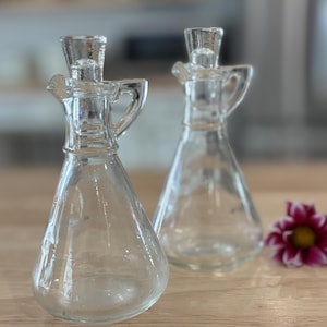 May include: Two clear glass cruets with conical bodies, small handles, and matching glass stoppers. Each vessel features a narrow neck and a wide base, designed for pouring liquids. The transparent glass construction allows for a clear view.
