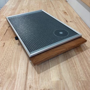May include: A rectangular, dark gray object with a silver frame and a wooden base. The top surface has a grid pattern and a circular design. The object is resting on a light-colored wooden surface.