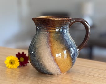 May include: A handmade ceramic pitcher with a dark brown rim and handle. The body of the pitcher features a blend of colors, including blue, white, and yellow-orange. Two flowers, one yellow and one red, are next to the pitcher.