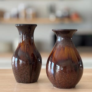 May include: Two brown ceramic vases with a glossy finish. The vases have a narrow neck and a wider base. The color transitions from a dark brown at the top to a lighter brown at the base. The vases are displayed on a wooden surface.