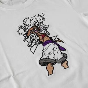 Puede incluir: Camiseta blanca con un personaje de dibujos animados bordado. El personaje tiene piel morena, pecho rojo y ropa blanca. El diseño incluye cabello negro y una faja morada. El diseño bordado está centrado en la camiseta.