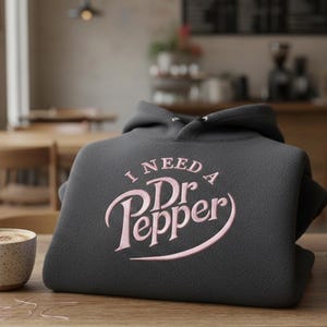 May include: A dark gray hoodie with the text "I NEED A Dr Pepper" in pink script. The hoodie is folded on a wooden table next to a cup of coffee and autumn leaves. The background is a blurred cafe setting.