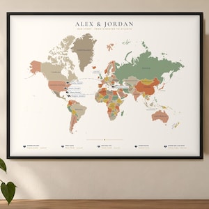 May include: A framed world map print titled ALEX & JORDAN OUR STORY - FROM KINGSTON TO ATLANTA. The map features muted green, tan, and orange landmasses on a cream background with dashed lines connecting specific locations marked by heart icons.