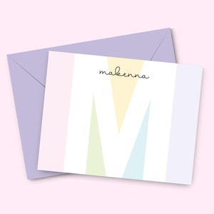 May include: A white card with a pastel colored geometric design and the name "Makenna" written in black script. The card is on top of a lavender envelope.