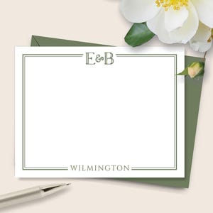 May include: A white stationery card with a green border, the initials "E&B" at the top, and the word "WILMINGTON" at the bottom. A silver pen rests near the card, and white flowers with yellow centers are in the background.