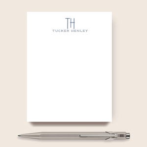 May include: A white notepad with the name "Tucker Henley" printed in navy blue, along with the initials "TH". A silver pen rests below the notepad, providing a sleek, modern aesthetic. The notepad and pen are set against a neutral background.