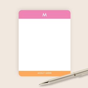 May include: A rectangular notepad with rounded corners. The top section is pink with a white "M". The bottom section is orange with the text "MOLLY ANNE". A silver pen rests on the side.
