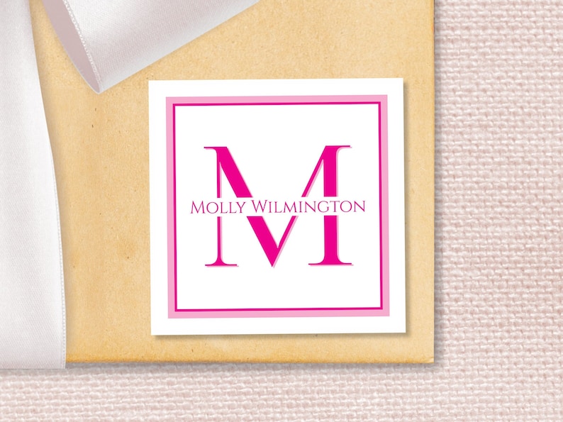 May include: A white square label with a pink border. The letter "M" is in a large pink font, with the text "MOLLY WILMINGTON" centered below the letter.