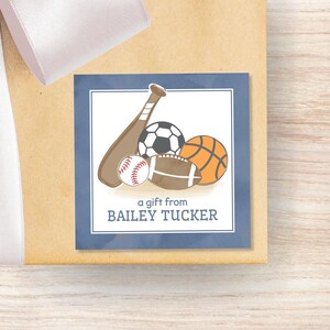 May include: A square gift tag with a dark blue border and a white center featuring illustrations of a brown baseball bat, a soccer ball, a white baseball, a brown football, and an orange basketball, with the text a gift from BAILEY TUCKER.
