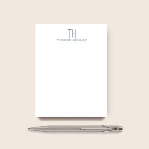 May include: A white rectangular notepad featuring the dark blue monogram TH and the name TUCKER HENLEY printed in a serif font at the top, accompanied by a metallic silver hexagonal mechanical pencil with a pocket clip positioned below the notepad.