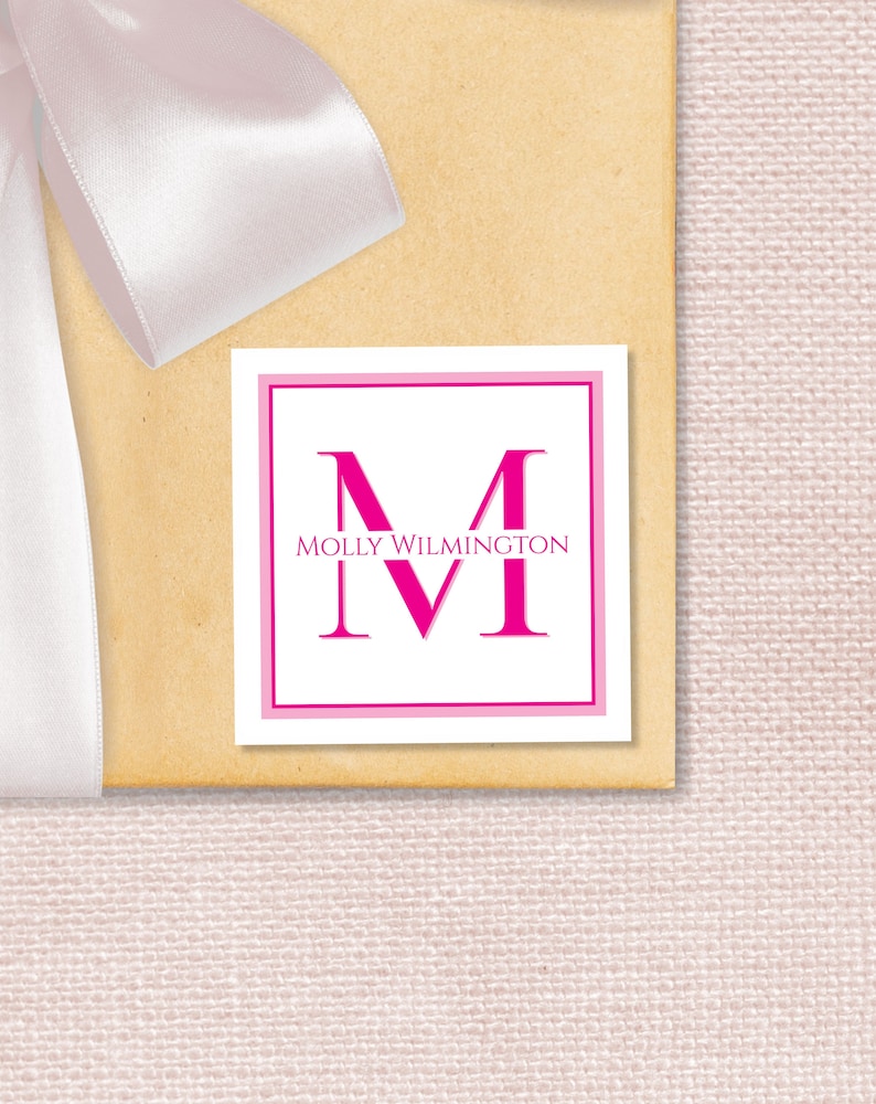 May include: A white square gift tag with a pink border. The letter "M" is in large, pink, bold font. The text "MOLLY WILMINGTON" is in smaller, black font below the letter.