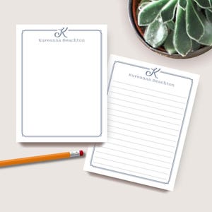 May include: Two white notepads with a blue border and the text "Kureanna Beachton". One notepad is blank, the other has lined paper. A yellow pencil with a red eraser is on the table. A potted succulent is in the background.