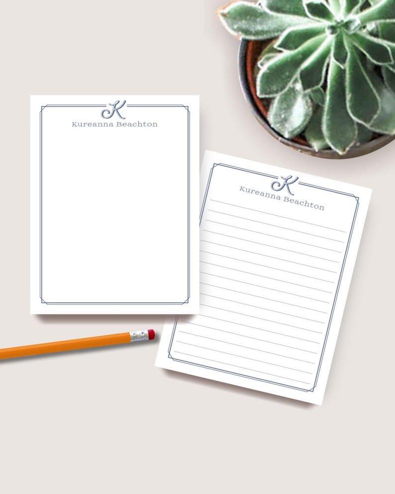 May include: Two white notepads with a blue border and the text "Kureanna Beachton". One notepad is blank, the other has lined paper. A yellow pencil with a red eraser is on the table. A potted succulent is in the background.