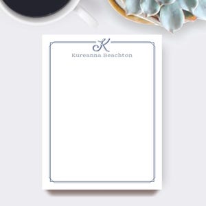 May include: A white notepad with a blue border and the name "Kureanna Beachton" printed in blue. The letter "K" is stylized at the top. A cup of coffee and a succulent plant are in the background.
