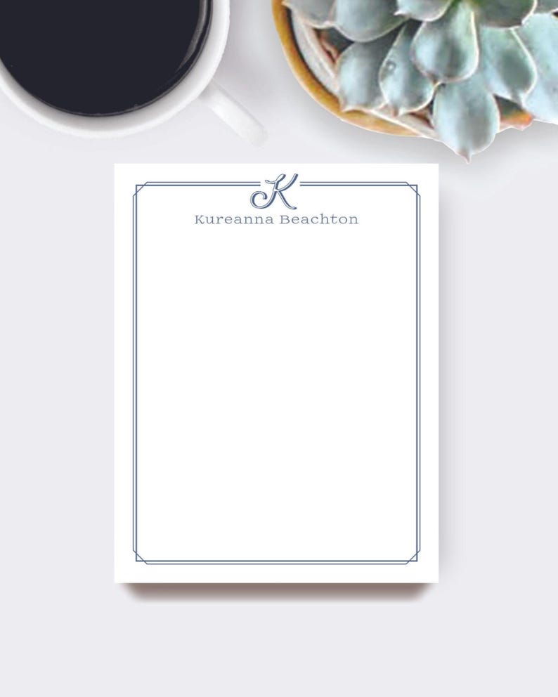 May include: A white notepad with a blue border and the name "Kureanna Beachton" printed in blue. The letter "K" is stylized at the top. A cup of coffee and a succulent plant are in the background.