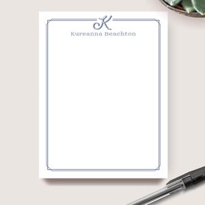 May include: A white notepad with a blue border and the name "Kureanna Beachton" printed in blue. A blue cursive "K" is above the name. A pen is in the bottom right corner. The notepad is on a light surface.