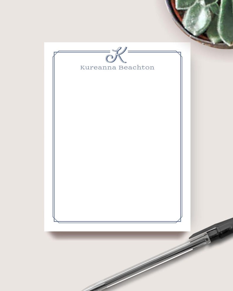 May include: A white notepad with a blue border and the name "Kureanna Beachton" printed in blue. A blue cursive "K" is above the name. A pen is in the bottom right corner. The notepad is on a light surface.
