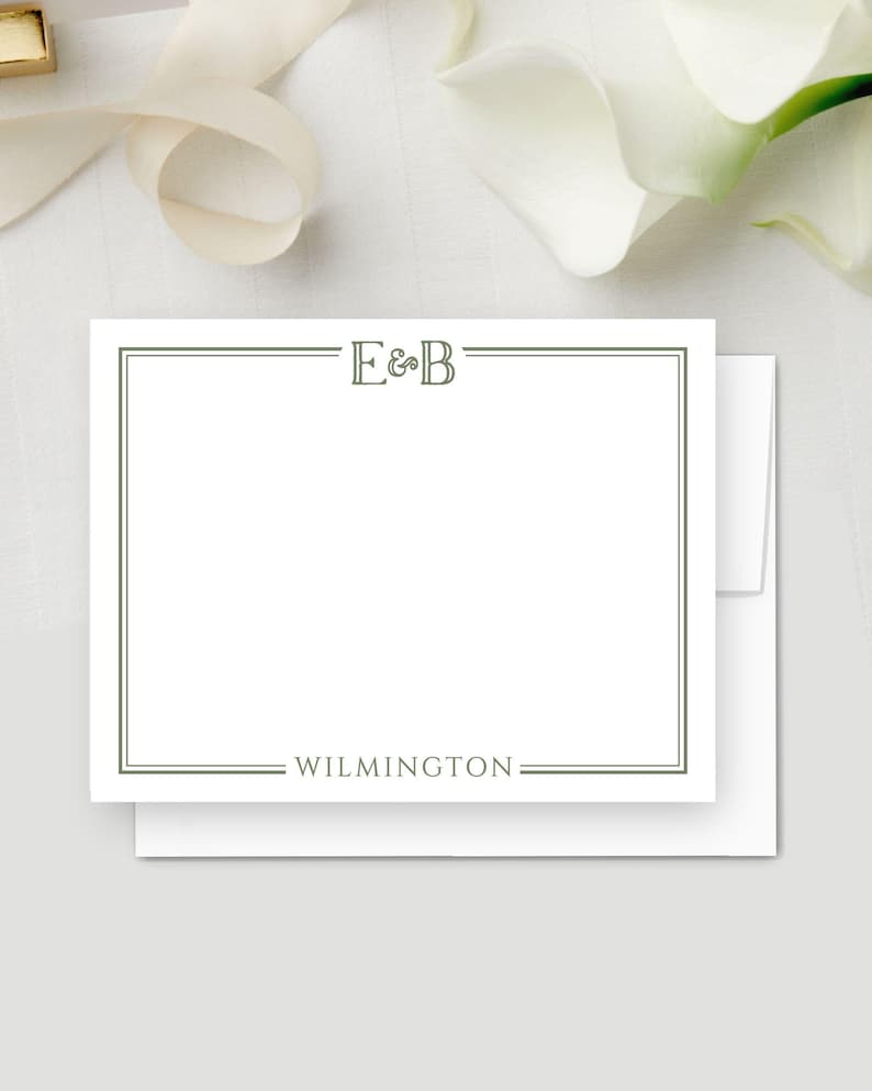 May include: White stationery card with a green border, the initials "E&B" at the top, and the word "WILMINGTON" at the bottom. The card is on top of a white envelope, with a white background and a ribbon.