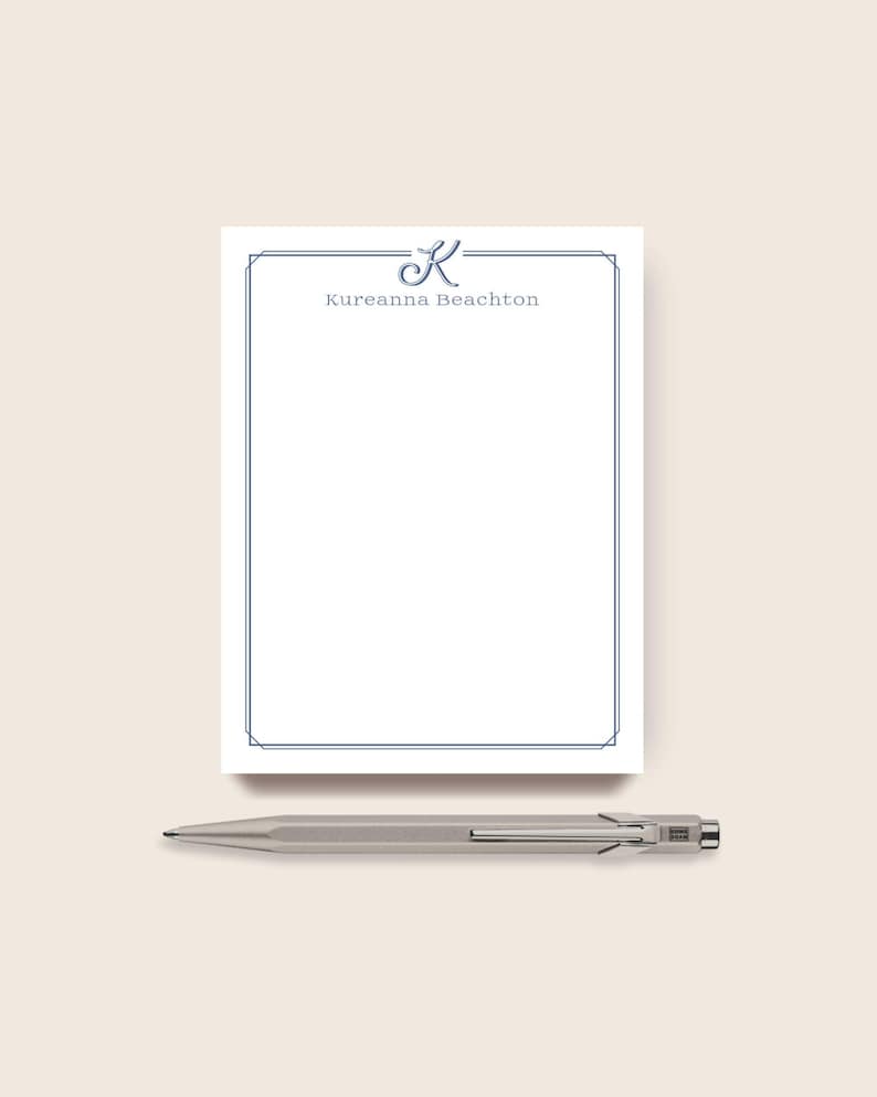 May include: A white notepad with a blue border and the name "Kureanna Beachton" printed in blue. A silver pen rests below the notepad. The notepad features a stylized letter "K" at the top.