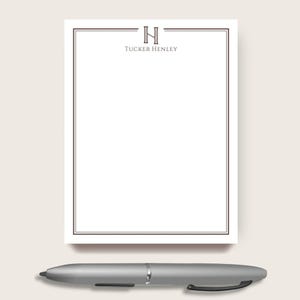 May include: A white rectangular notepad featuring a dark brown double-line border and a centered dark brown letter H monogram above the text Tucker Henley. A silver-toned ballpoint pen with a black clip is positioned horizontally below the notepad.