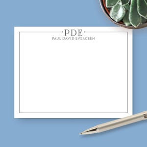 May include: A rectangular white notepad featuring a thin black border and centered text that reads PDE and PAUL DAVID EVERGEEN in a serif font. The notepad is positioned on a solid light blue surface with a small succulent and a silver pen.