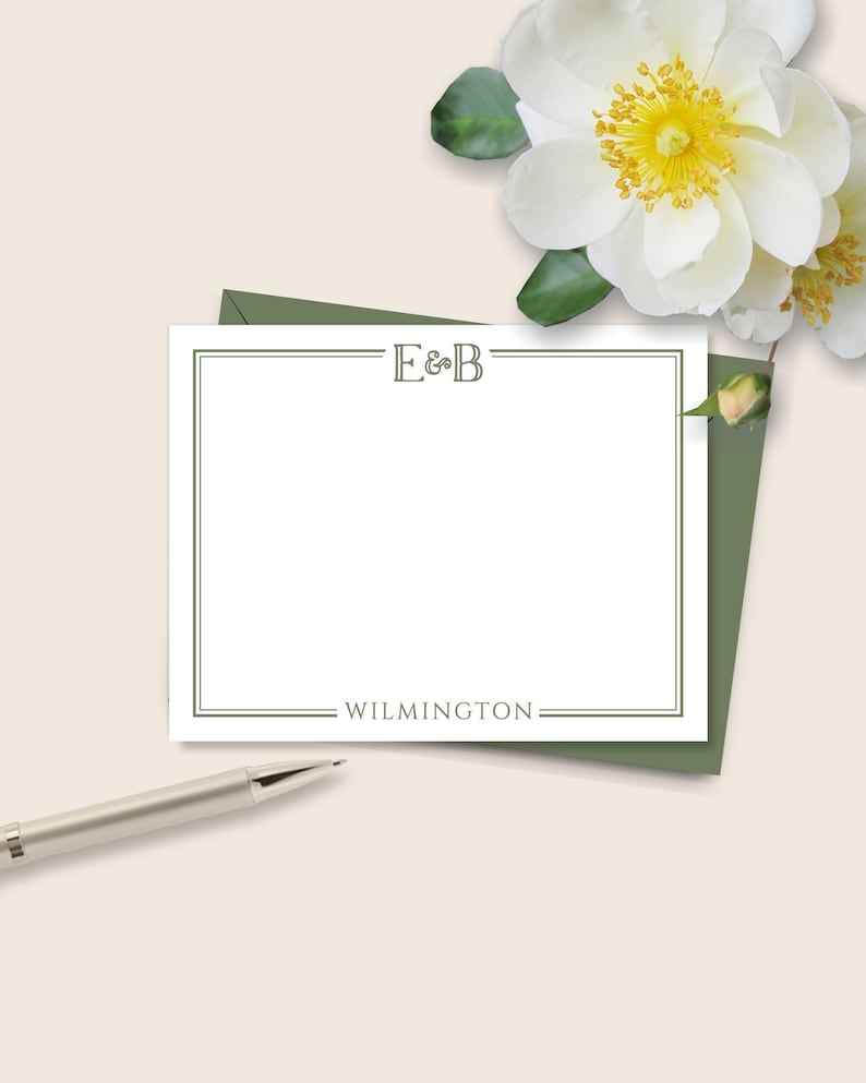 May include: A white stationery card with a green border, the initials "E&B" at the top, and the word "WILMINGTON" at the bottom. A silver pen rests near the card, and white flowers with yellow centers are in the background.