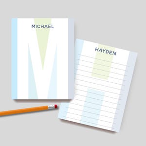 May include: Two personalized notepads with the names "MICHAEL" and "HAYDEN" printed in a dark blue font. The "MICHAEL" notepad features a large "M" design in light blue and green. A yellow pencil with a red eraser rests nearby.
