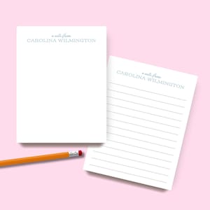 May include: Two white rectangular stationery sheets featuring the printed text a note from CAROLINA WILMINGTON in grey ink. One sheet is blank, while the other contains horizontal grey lines. An orange pencil with a silver ferrule is included.