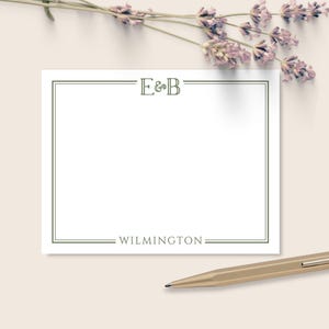 May include: A white stationery card with a green border, the initials "E&B" at the top, and the word "WILMINGTON" at the bottom. A gold pen rests on the surface, with lavender sprigs in the background.