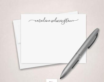 May include: A white rectangular stationery card featuring the black cursive text caroline wilmingtons centered at the top, layered over a matching white envelope, accompanied by a silver ballpoint pen positioned diagonally across the card.