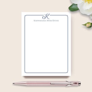 May include: A white notepad with a blue border and the name "Kureanna Beachton" printed in blue. A rose gold pen rests below the notepad. A white flower with a yellow center is in the background.