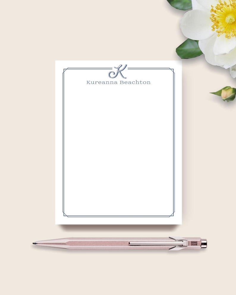 May include: A white notepad with a blue border and the name "Kureanna Beachton" printed in blue. A rose gold pen rests below the notepad. A white flower with a yellow center is in the background.