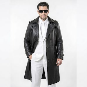 Pode incluir: Um trench coat longo de couro preto com design de abotoamento duplo. O casaco é usado sobre um fato branco, criando um contraste marcante. O modelo usa óculos de sol pretos. O casaco tem cerca de 130 cm de comprimento.