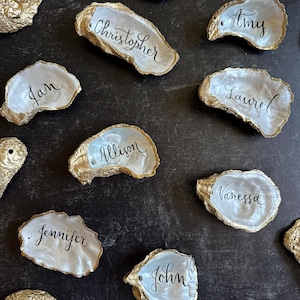 May include: Gold-painted oyster shells with handwritten names in black script. Names include Christopher, Amy, Jan, Laurel, Allison, Jennifer, John, and Vanessa. The shells are arranged on a dark surface, suitable for place settings.
