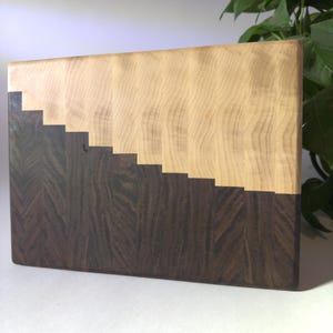 May include: A wooden cutting board with a light wood top and a dark wood bottom. The two woods are separated by a stepped pattern.