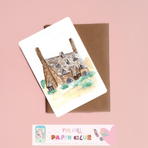 Peut inclure: Illustration à l'aquarelle d'une maison en pierre avec un toit en ardoise et deux cheminées. L'image est sur une carte blanche, avec une enveloppe marron derrière. La carte porte le texte "FAN GIRL PAPER CLUB" en bas.